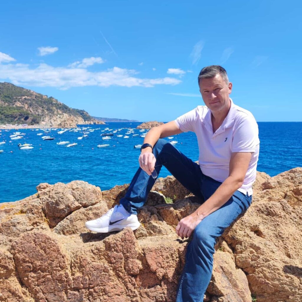 Sascha Begoll sitzt entspannt auf Felsen an einer mediterranen Küste mit blauem Meer im Hintergrund.